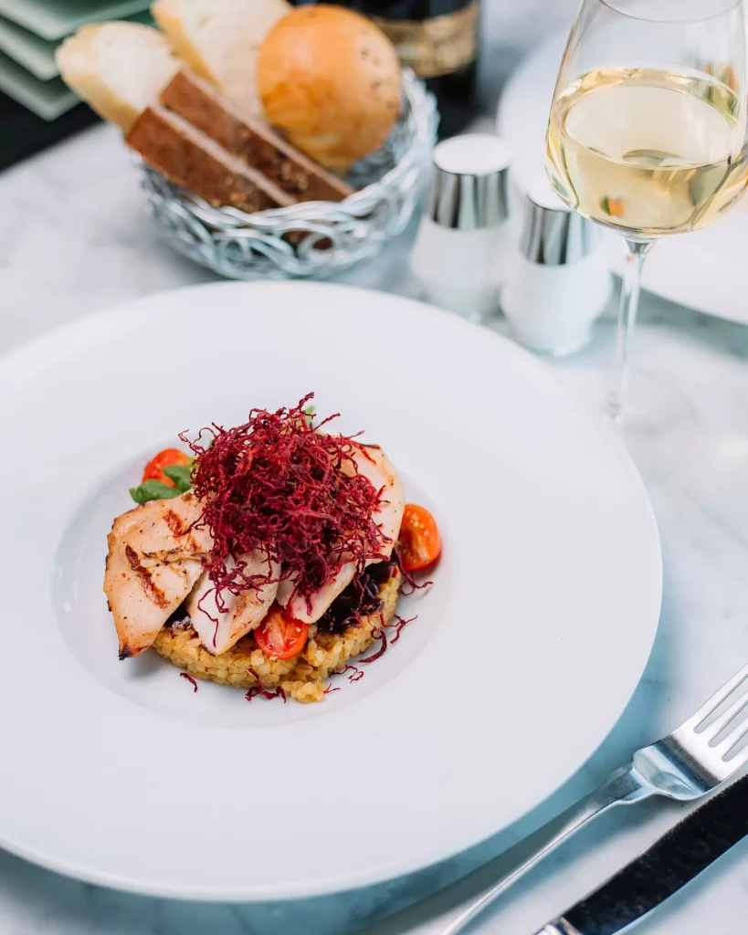 Plat de poulet grillé avec riz, tomates cerises et garniture rouge, verre de vin blanc et panier de pain sur table en marbre.