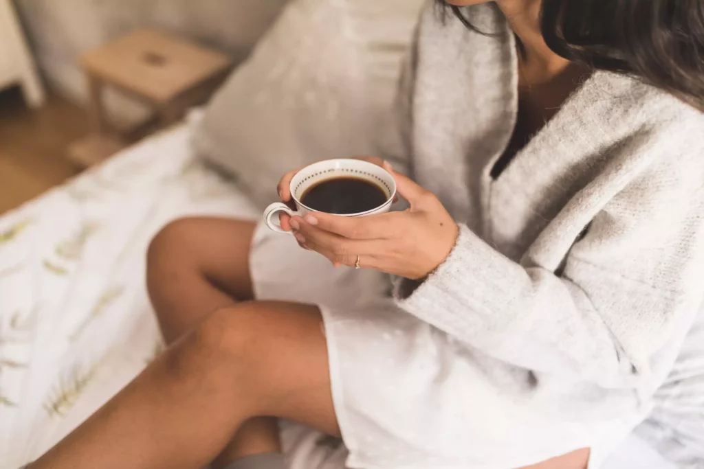 Woman in a cosy jumper sitting on a bed and holding a cup of coffee in an apartment at Maison Talmühle Black Forest Resort