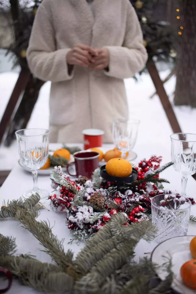 Winter table decoration with fir branches, red berries, mandarins and glasses before a snowy Black Forest backdrop